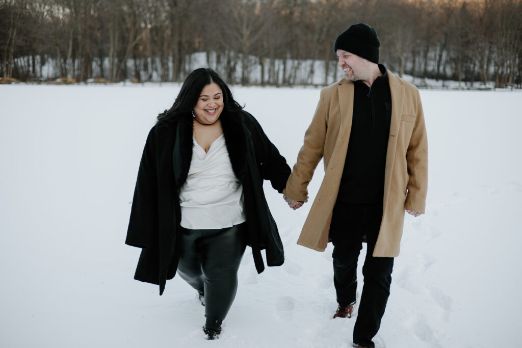 Couple smiling and holding hands, walking through the snow towards the camera in Connecticut