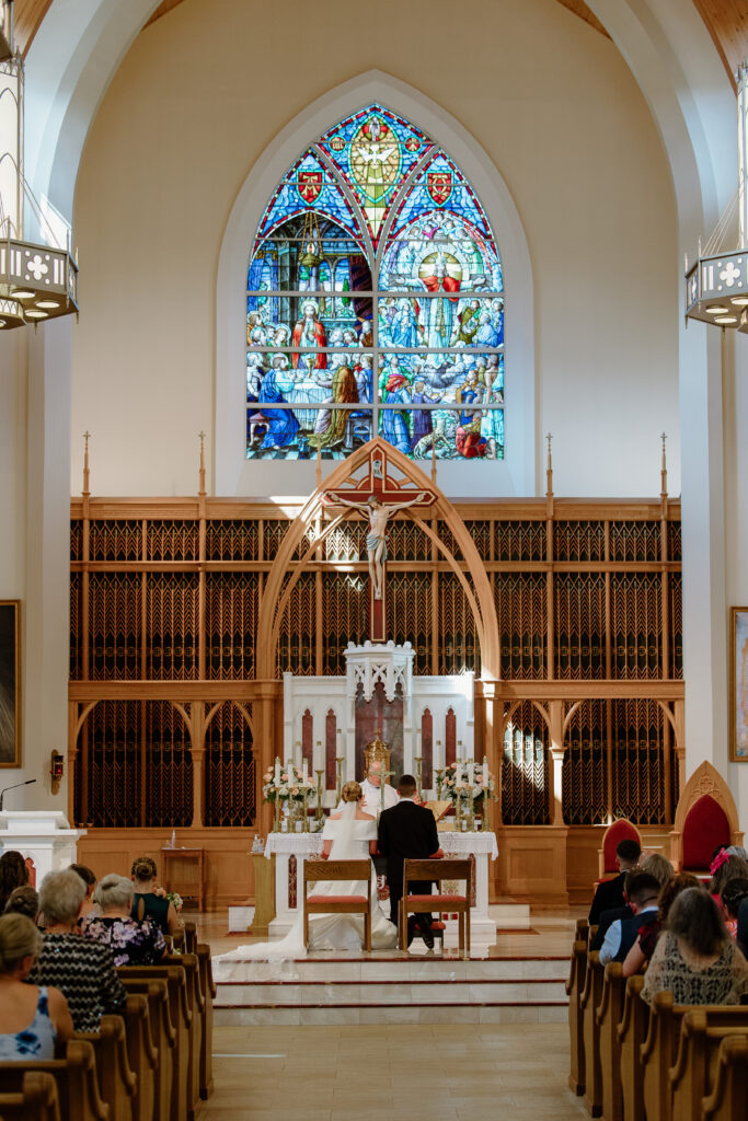 Wide view of Clara and Matt seated together during their church ceremony, surrounded by stained glass and guests, capturing a quiet moment from their Curry Estate wedding day.