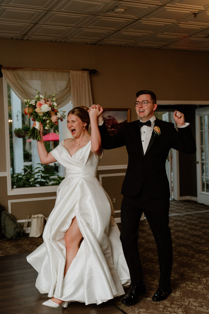 Clara and Matt celebrate as they enter their reception, smiling and raising their arms together during their Curry Estate wedding.