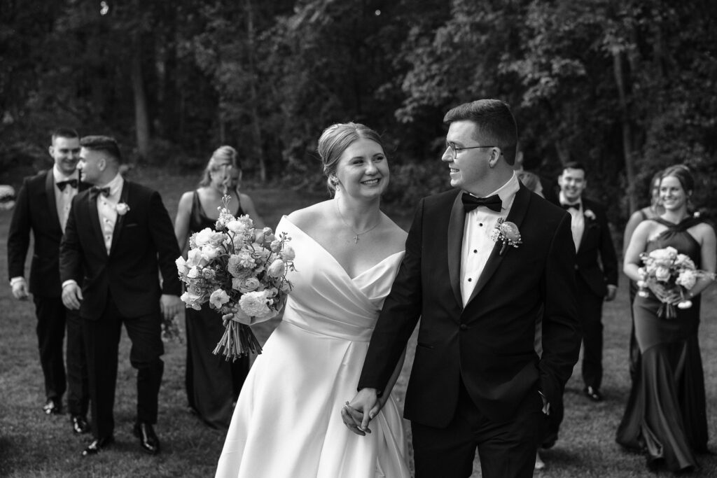 Clara and Matt walk hand in hand after their ceremony, smiling at each other as their wedding party follows behind during their Curry Estate wedding.