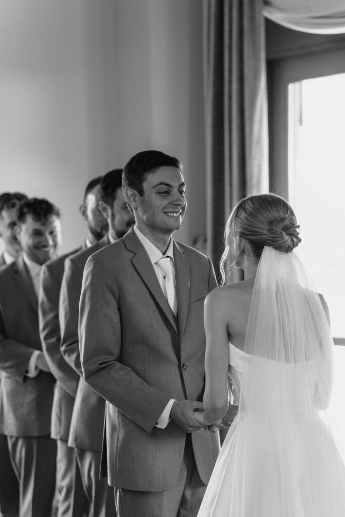 Black and white photo of the groom smiling at his bride as they hold hands at the altar