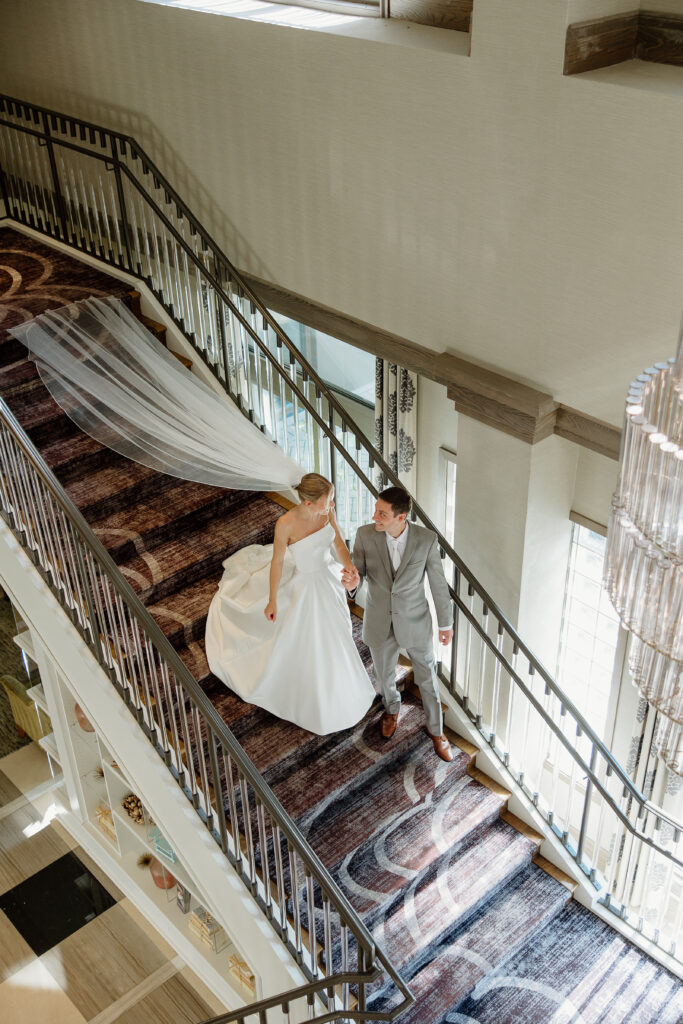 Overhead view of a bride and groom walking down a grand staircase, her veil floating behind her