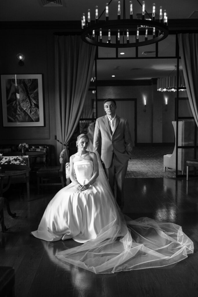 Black and white editorial portrait of a seated bride with her veil pooled on the floor and the groom standing behind her