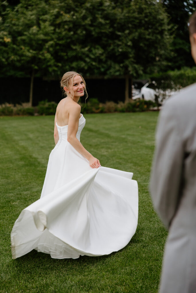 Bride twirling her satin skirt on the lawn and smiling back at the groom