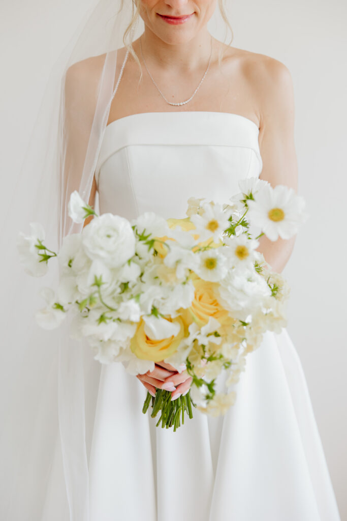 Close-up of a bride holding a bouquet of white sweet peas, ranunculus, daisies, and butter yellow roses