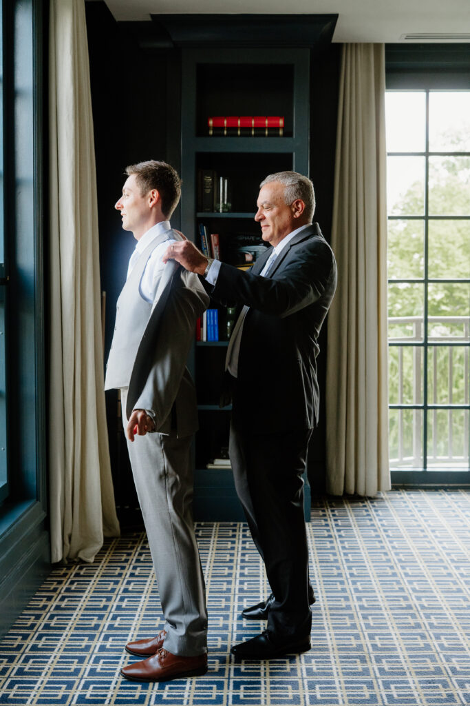 Groom getting help putting on his grey suit jacket beside a tall window