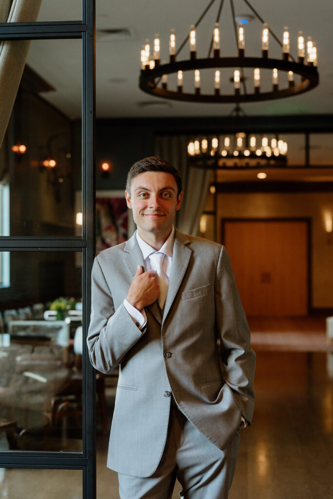 Groom in a light grey suit adjusting his tie under a ring chandelier at The Delamar West Hartford