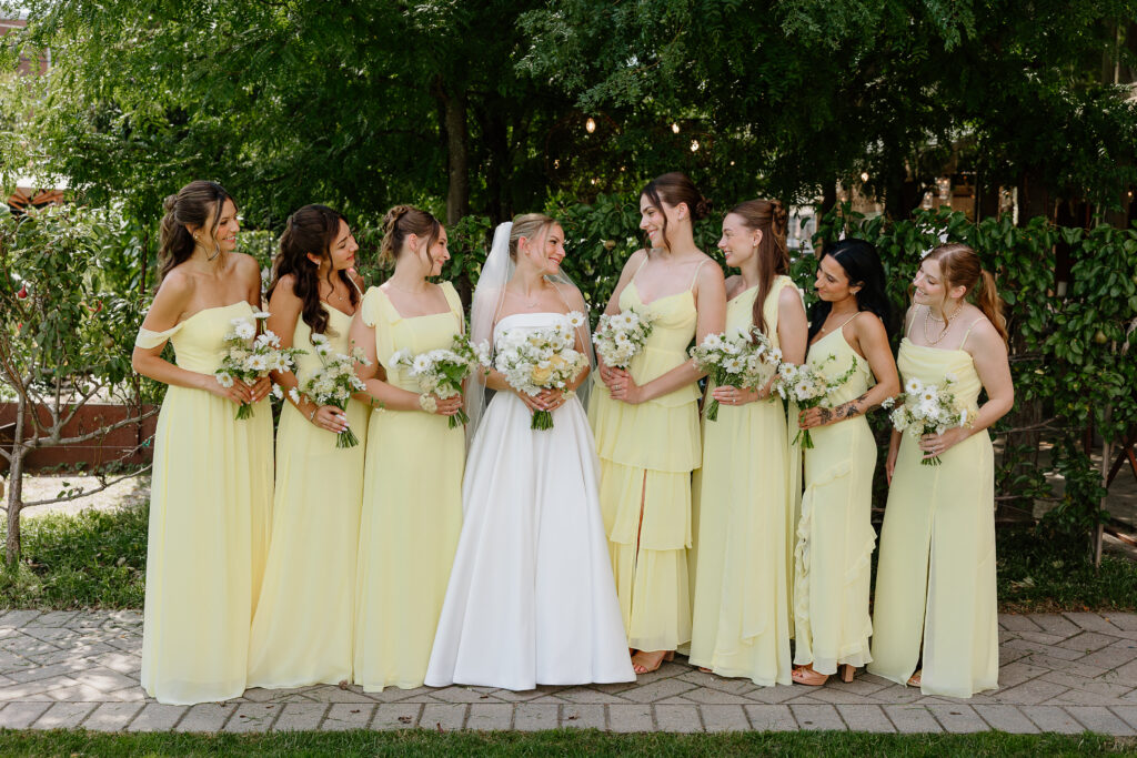 Bride with seven bridesmaids in mismatched butter yellow chiffon dresses holding white bouquets