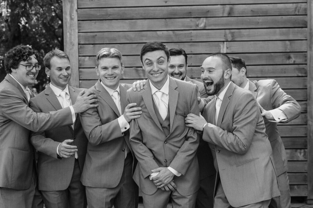 Black and white photo of groomsmen in grey suits laughing and crowding around the groom