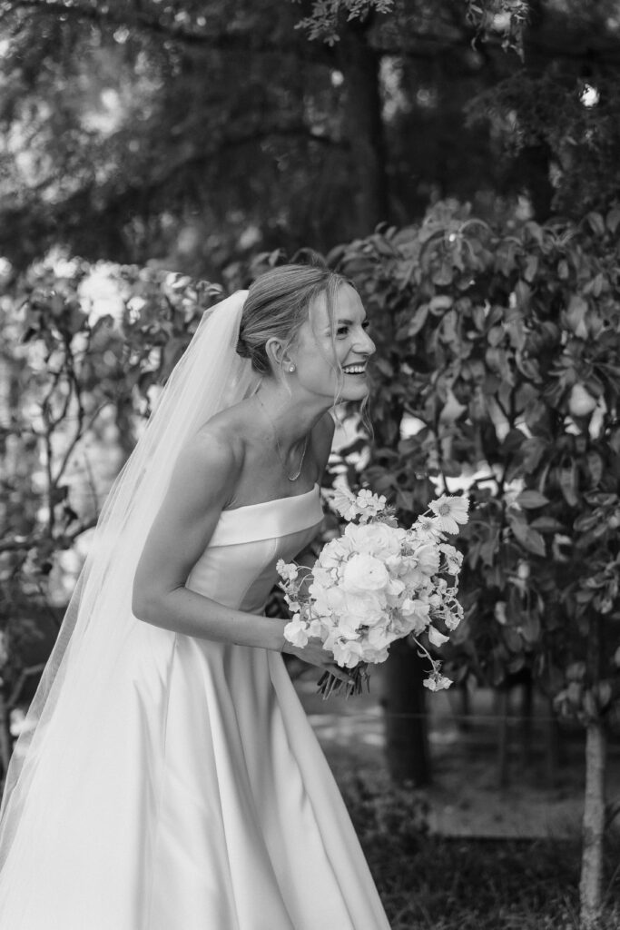 Black and white photo of a bride grinning and leaning forward with her bouquet before her father's first look