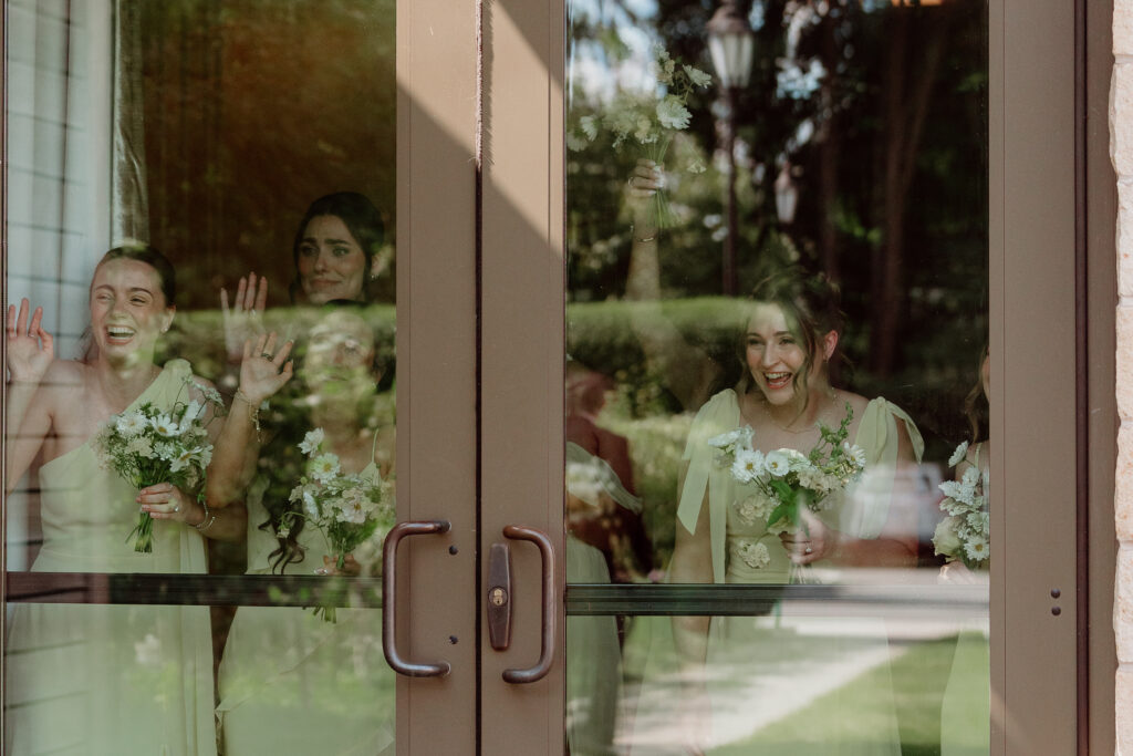 Bridesmaids in butter yellow peeking through glass doors to watch the father-daughter first look