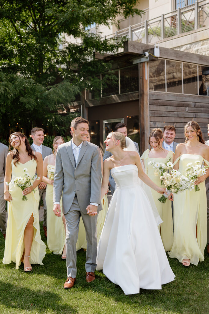 Bride and groom laughing as they walk ahead of bridesmaids in butter yellow dresses and groomsmen in grey suits